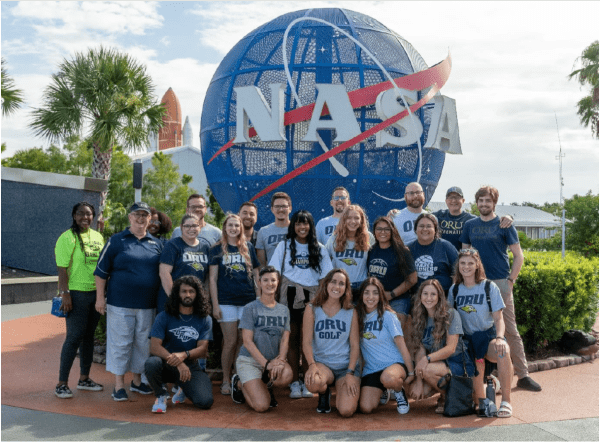 A group of ORU students smiling with ORU t-shirts in front of a NASA globe